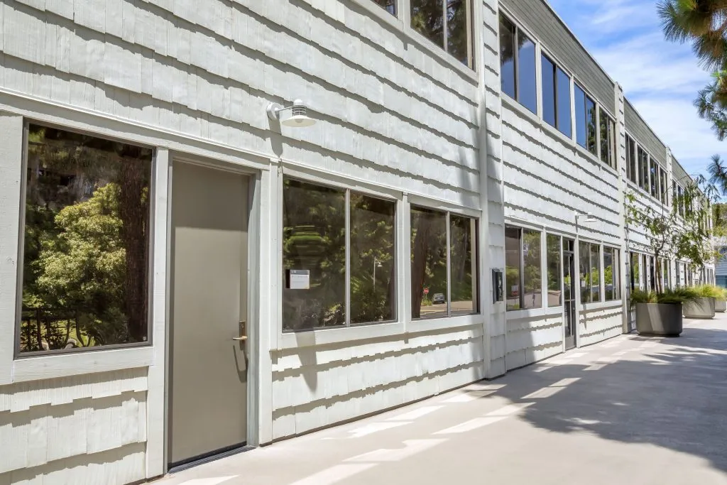 Two-story commercial building with white horizontal siding, multiple windows, and concrete walkway; trees visible left