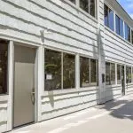 Two-story commercial building with white horizontal siding, multiple windows, and concrete walkway; trees visible left