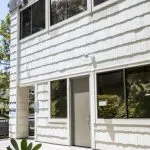 Modern two-story structure with white paneling, large reflective windows, glass entrance, and green foliage at base