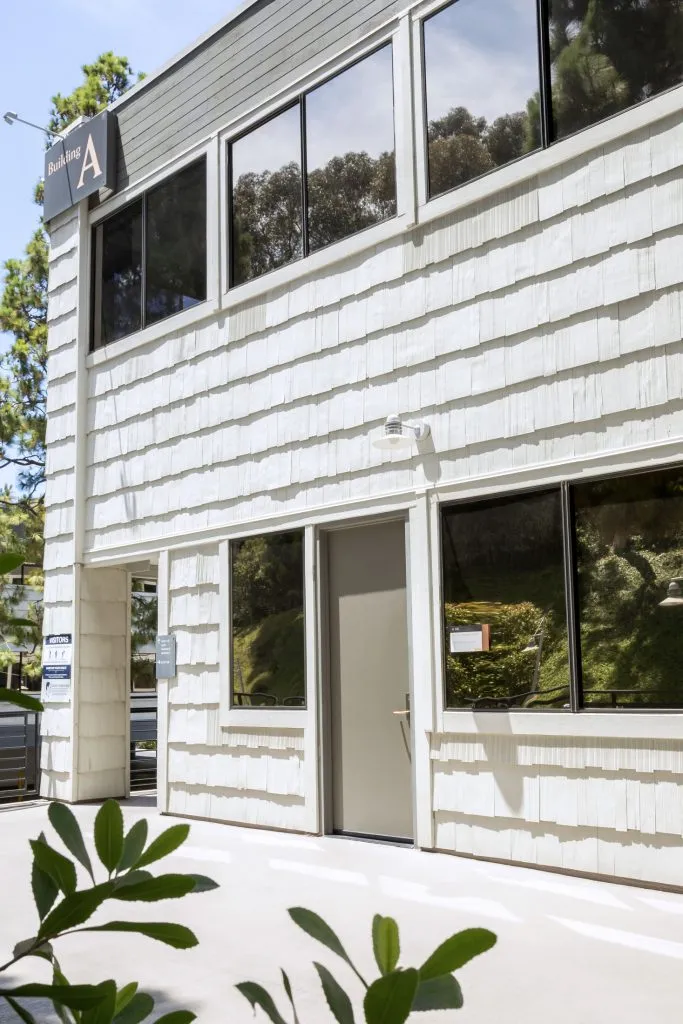 Modern two-story structure with white paneling, large reflective windows, glass entrance, and green foliage at base
