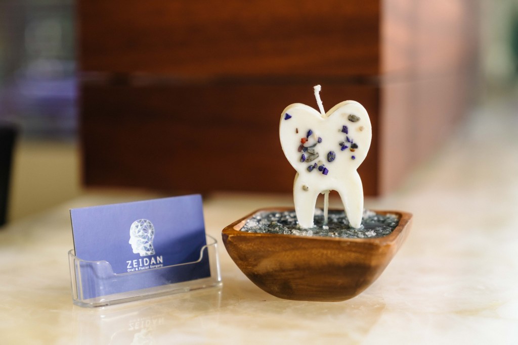 Decorative tooth-shaped candle with embedded colored specks, placed in wooden bowl on countertop; beside it, a clear holder with a “ZELDAN” business card