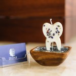 Decorative tooth-shaped candle with embedded colored specks, placed in wooden bowl on countertop; beside it, a clear holder with a “ZELDAN” business card