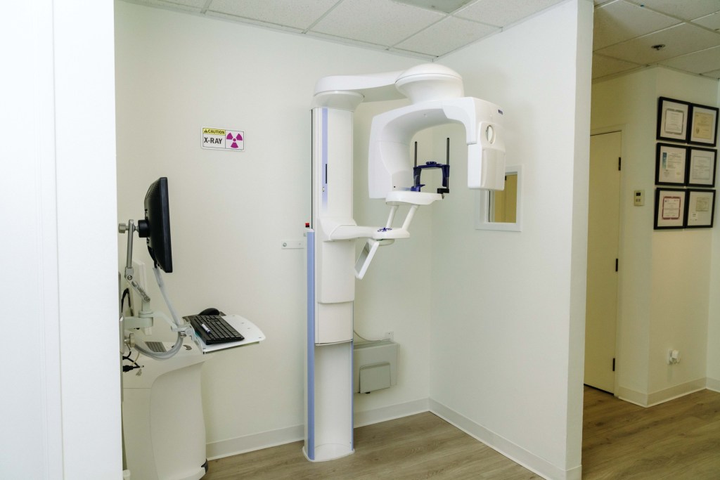 Panoramic X-ray unit mounted on ceiling track in white-walled room; monitor and control panel visible, door open to hallway