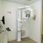 Panoramic X-ray unit mounted on ceiling track in white-walled room; monitor and control panel visible, door open to hallway