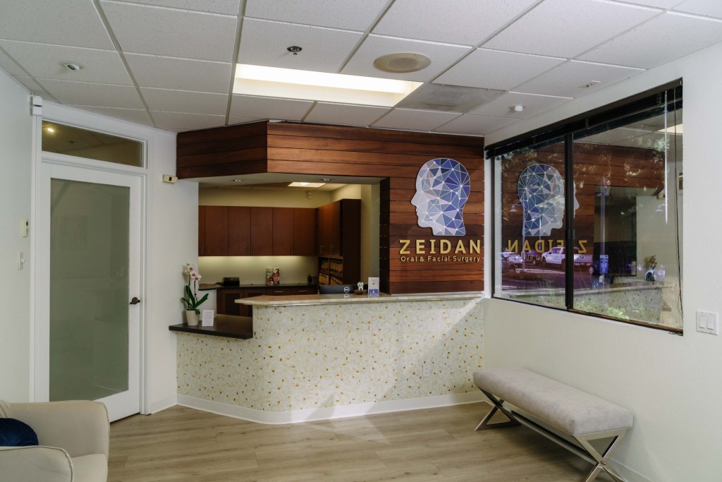 Reception area: white counter, wooden cabinet, framed artwork, and glass partition; door with green sign on left
