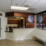 Reception area: white counter, wooden cabinet, framed artwork, and glass partition; door with green sign on left