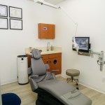 Operatory with gray dental chair, orange cabinetry, computer monitor, and wall-mounted equipment; no people present