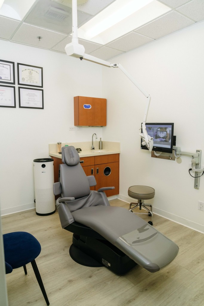 Operatory with gray dental chair, orange cabinetry, computer monitor, and wall-mounted equipment; no people present
