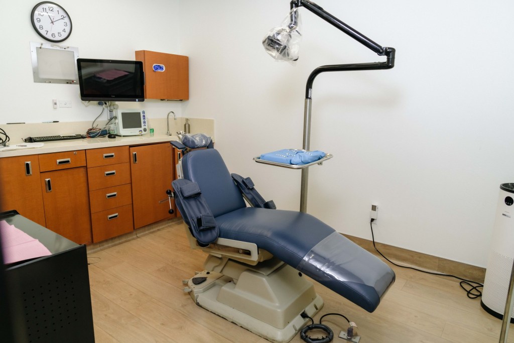 Operatory with blue dental chair, adjustable overhead light, orange cabinetry, wall-mounted monitor, and beige flooring; no people visible