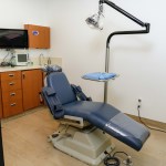 Operatory with blue dental chair, adjustable overhead light, orange cabinetry, wall-mounted monitor, and beige flooring; no people visible
