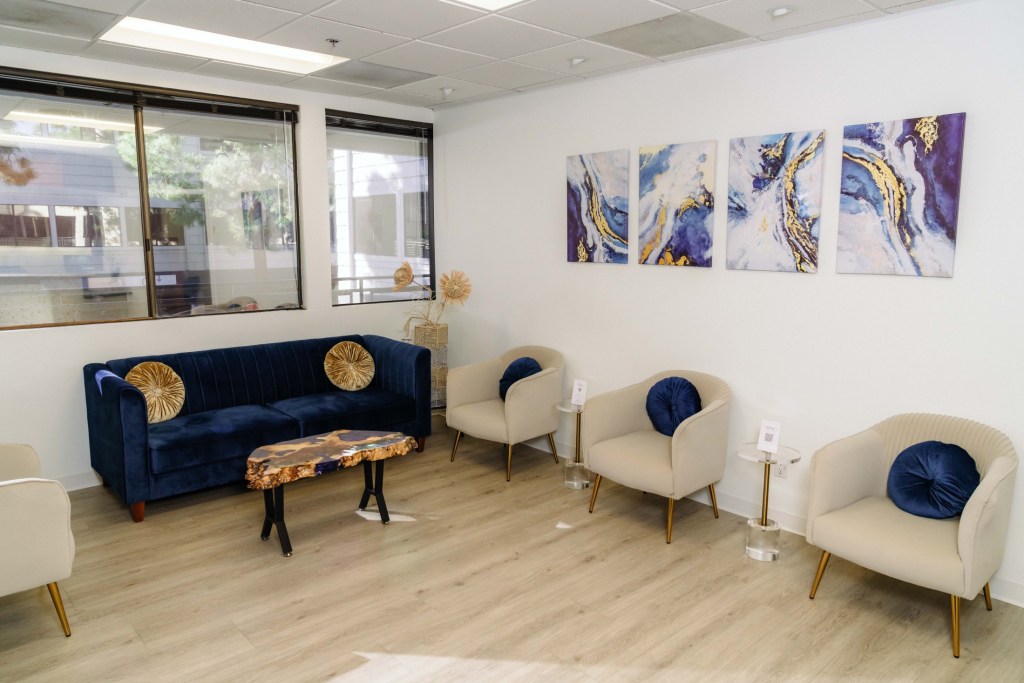 Waiting room with blue sofa, three beige armchairs, framed abstract art, wood-look flooring, and large windows