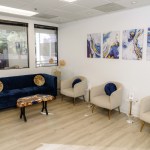 Waiting room with blue sofa, three beige armchairs, framed abstract art, wood-look flooring, and large windows